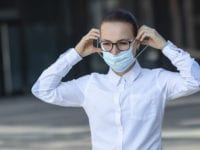 Beautiful business woman, young girl putting on medical protective mask on her face, in white shirt in glasses outdoors, healthcare at job, work, office. Coronavirus, virus, epidemic, covid-19 concept